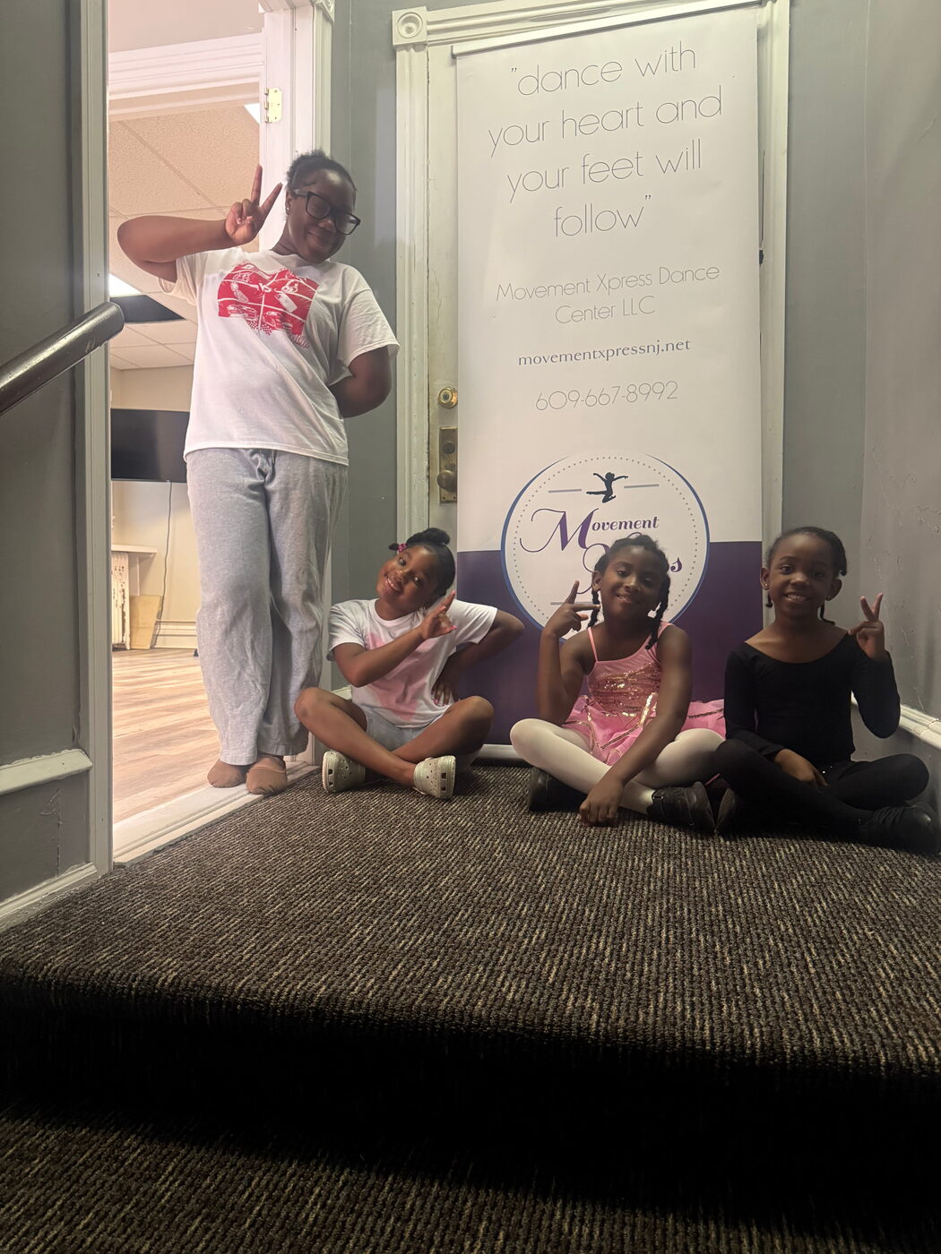 An adult and three young dancers smiling and making peace signs in front of a Movement Xpress banner